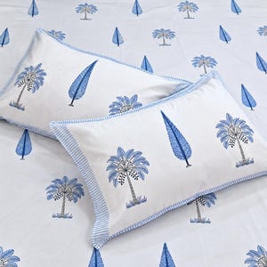 Può includere: Un lenzuolo bianco e due federe rettangolari abbinate con un motivo ripetuto di palme blu e forme di foglie blu. Le federe presentano un bordo a righe azzurre e bianche lungo i margini del tessuto.