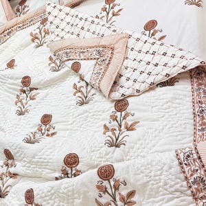May include: White quilted bedding set with a floral pattern in shades of brown and green. The set includes a quilt and pillowcases, featuring a repeating flower design and striped borders. The bedding is likely made of a soft, comfortable material.