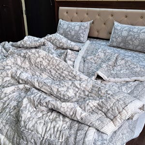 May include: A grey and white floral quilt set on a bed. The set includes a quilt, two pillowcases, and a bed sheet. The bedding features a detailed floral design and is made of a soft material. The headboard is beige with a button-tufted design.