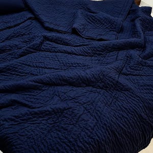 May include: A deep navy blue quilt featuring a textured, quilted surface with a repeating stitched pattern. The fabric is draped across a bed, showing folds and creases throughout the material, creating a soft and layered appearance.