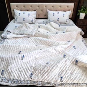 May include: A white bedspread and pillow set with a blue floral pattern. The bedding is arranged on a bed with a tufted headboard. The quilt has a quilted design and a patterned border.