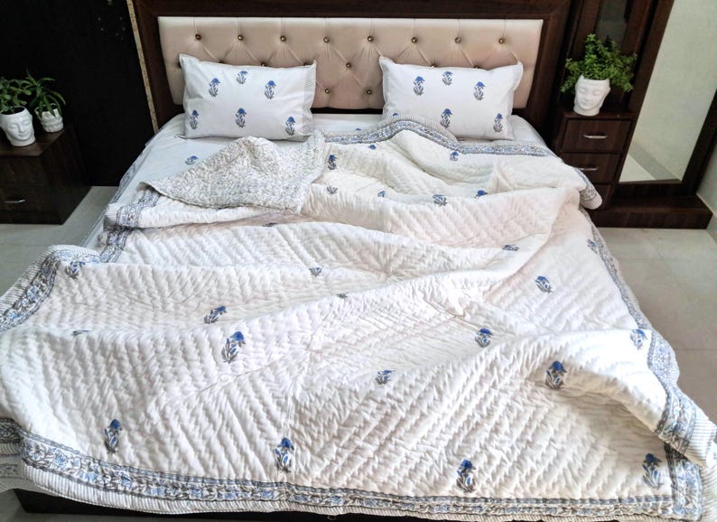 May include: A white bedspread and pillow set with a blue floral pattern. The bedding is arranged on a bed with a tufted headboard. The quilt has a quilted design and a patterned border.