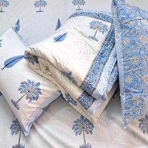 May include: A bedding set featuring a white quilt and two pillows with blue palm tree and leaf patterns. The quilt has a blue floral border. The pillow on the right has a blue floral pattern. The bedsheet also has a blue palm tree pattern.