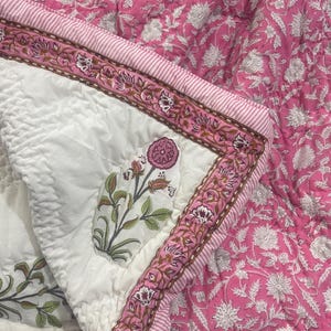 May include: A pink floral quilt with a white center and a decorative border. The quilt features a floral pattern in shades of pink and white. The border has a striped design and a floral pattern.
