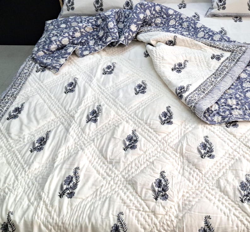 Puede incluir: Colcha de algod&oacute;n blanco con un patr&oacute;n de diamante y estampado floral negro. La colcha est&aacute; doblada por la mitad y tiene un estampado floral azul y blanco en el reverso.