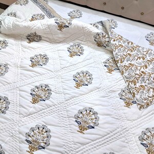 Puede incluir: Colcha acolchada blanca con un estampado floral repetido en tonos grises, azules y dorados. La colcha está sobre una cama con una sábana blanca y una funda de almohada estampada. El tema general es floral.
