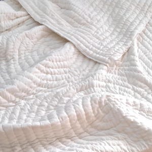 May include: A white, quilted blanket with a textured surface. The blanket has a repeating circular pattern and a folded edge. The fabric appears soft and comfortable, suitable for bedding or home decor.