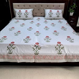 Könnte beinhalten: Weißes Bettwäsche-Set mit Blumenmuster und zwei Kissenbezügen. Die Bettwäsche zeigt ein sich wiederholendes Muster aus roten Blumen und grünen Blättern. Der Rand des Lakens und der Kissenbezüge hat ein rosa-graues Blumenmuster.