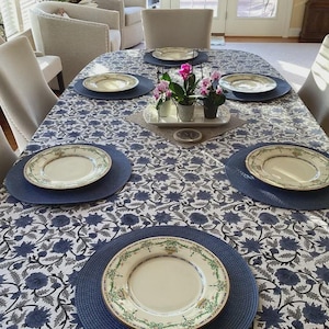 Könnte beinhalten: Ein ovaler Esstisch, gedeckt für eine Mahlzeit. Der Tisch ist mit einer blau-weißen Blumendecke bedeckt. Jedes Gedeck umfasst einen blauen Platzdeckchen und einen dekorativen Teller. In der Mitte befindet sich ein kleines Blumengesteck.