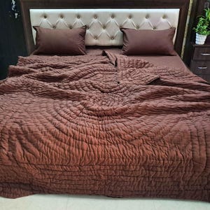 Op de afbeelding: Een bed met een bruin gewatteerd dekbed en bijpassende kussenslopen. Het hoofdbord is bekleed met een lichtbeige stof met een knoop-getuft ontwerp. Twee nachtkastjes met planten zijn zichtbaar.