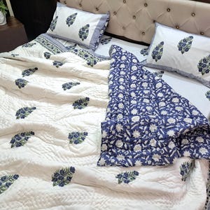 Könnte beinhalten: Eine weiße, gesteppte Tagesdecke und Kissenbezüge mit blauem Blumenmuster. Das Bett ist mit einer blau-weiß gemusterten Decke bezogen. Das Bettwäsche-Set befindet sich auf einem Bett mit beigem Kopfteil.