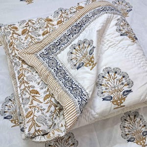 May include: A folded white quilt with a floral pattern in gold, gray, and blue. The quilt has a striped border and a repeating floral design on the white background. The quilt is likely made of cotton.