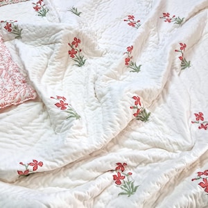Op de afbeelding: Witte gewatteerde beddengoedset met een bloemenpatroon in rood en groen. De set bevat een quilt en een kussen met een bijpassend ontwerp. De quilt heeft een herhalend patroon van rode bloemen en groene bladeren.