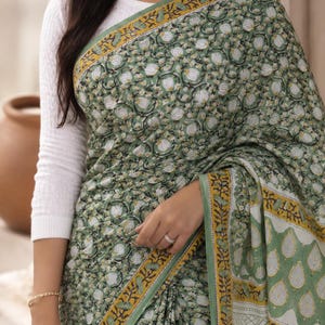 May include: A sage green saree featuring a dense floral block print pattern in white and dark green, accented by a mustard yellow and green geometric border. The garment is paired with a long-sleeved white textured blouse with a round neckline.