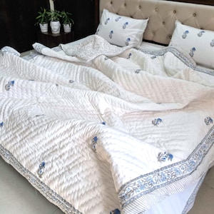 May include: White quilted bedding set with blue floral accents. The set includes a comforter and two pillowcases. The comforter has a detailed border and a repeating floral pattern. The bedding is on a bed with a beige headboard.
