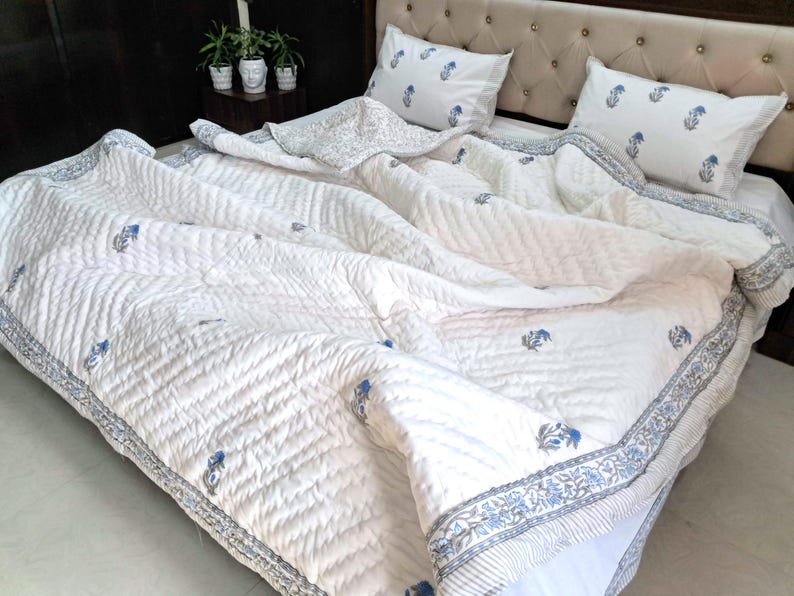 May include: White quilted bedding set with blue floral accents. The set includes a comforter and two pillowcases. The comforter has a detailed border and a repeating floral pattern. The bedding is on a bed with a beige headboard.