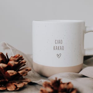 Peut inclure: Une tasse en céramique blanche avec une finition mouchetée et un bord brun. La tasse porte l'inscription "Ciao Kakao" et un petit cœur imprimé sur le devant.