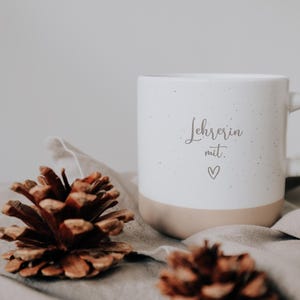 Weiße Keramiktasse mit der Aufschrift "Lehrerin mit Herz" auf einem weichen Stoff, umgeben von Tannenzapfen. Ideal als Geschenk für Lehrerinnen, die mit Leidenschaft und Liebe unterrichten. Diese stilvolle Tasse verbindet Funktionalität mit einer persönlichen Note und bereichert jeden Morgenkaffee oder Nachmittagstee. Perfekt, um Wertschätzung und Dankbarkeit auszudrücken.