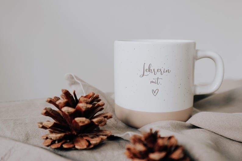 Weiße Keramiktasse mit der Aufschrift "Lehrerin mit Herz" auf einem weichen Stoff, umgeben von Tannenzapfen. Ideal als Geschenk für Lehrerinnen, die mit Leidenschaft und Liebe unterrichten. Diese stilvolle Tasse verbindet Funktionalität mit einer persönlichen Note und bereichert jeden Morgenkaffee oder Nachmittagstee. Perfekt, um Wertschätzung und Dankbarkeit auszudrücken.