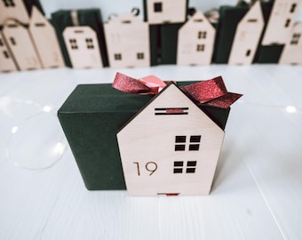 May include: A wooden house-shaped box with the number 19 on the front, part of an advent calendar. The box is light brown with four square windows and a red ribbon. The box is on a white surface.