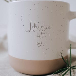 Weiße Tasse mit der Aufschrift "Lehrerin mit Herz" auf einem strukturierten Tuch platziert, umgeben von zarten, grünen Zweigen. Diese elegante und liebevoll gestaltete Tasse ist das perfekte Geschenk für Lehrerinnen, die mit Leidenschaft und Hingabe unterrichten. Ideal für den täglichen Kaffeegenuss und als Zeichen der Wertschätzung.