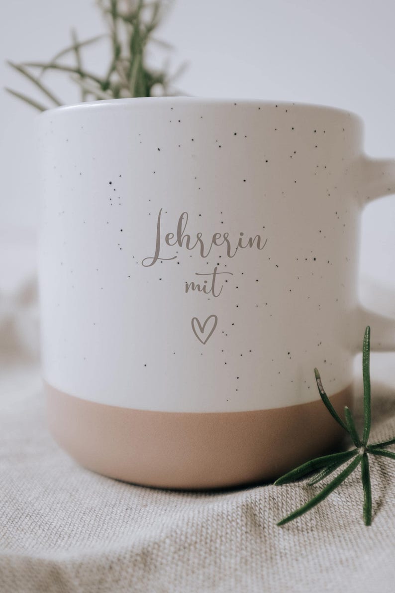Weiße Tasse mit der Aufschrift "Lehrerin mit Herz" auf einem strukturierten Tuch platziert, umgeben von zarten, grünen Zweigen. Diese elegante und liebevoll gestaltete Tasse ist das perfekte Geschenk für Lehrerinnen, die mit Leidenschaft und Hingabe unterrichten. Ideal für den täglichen Kaffeegenuss und als Zeichen der Wertschätzung.