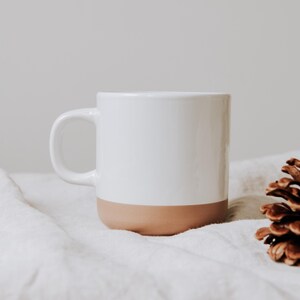 Elegante Keramiktasse mit schlichtem Design, ideal für gemütliche Stunden. Die Kombination aus weißem und beigefarbenem Ton sorgt für eine warme, einladende Atmosphäre. Perfekt für Ihren Morgenkaffee oder Tee am Nachmittag. Die Tasse steht auf weichem Stoff, flankiert von Tannenzapfen, und verleiht ein Gefühl von Geborgenheit und Naturverbundenheit.