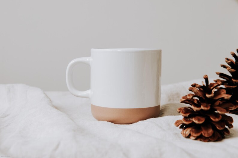 Elegante Keramiktasse mit schlichtem Design, ideal für gemütliche Stunden. Die Kombination aus weißem und beigefarbenem Ton sorgt für eine warme, einladende Atmosphäre. Perfekt für Ihren Morgenkaffee oder Tee am Nachmittag. Die Tasse steht auf weichem Stoff, flankiert von Tannenzapfen, und verleiht ein Gefühl von Geborgenheit und Naturverbundenheit.