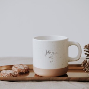 Elegante Tasse mit der Aufschrift "Lehrerin mit Herz" auf einem goldenen Tablett, umgeben von Keksen und Tannenzapfen. Perfekt für gemütliche Momente und ein ideales Geschenk für engagierte Lehrerinnen. Die warme Farbpalette und das liebevolle Design wecken ein Gefühl von Geborgenheit und Dankbarkeit.
