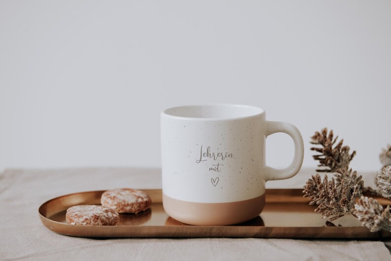 Elegante Tasse mit der Aufschrift "Lehrerin mit Herz" auf einem goldenen Tablett, umgeben von Keksen und Tannenzapfen. Perfekt für gemütliche Momente und ein ideales Geschenk für engagierte Lehrerinnen. Die warme Farbpalette und das liebevolle Design wecken ein Gefühl von Geborgenheit und Dankbarkeit.