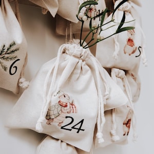 Puede incluir: Calendario de Adviento con bolsas de cordón de color crema. Cada bolsa está numerada y decorada con ilustraciones de animales y vegetación festiva. Las bolsas cuelgan sobre un fondo blanco.