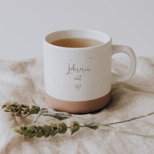 Handgefertigte Keramiktasse mit der Aufschrift "Lehrerin mit Herz" auf einem beigen Leinentuch, umgeben von zarten, getrockneten Blumen. Diese einzigartige Tasse ist das perfekte Geschenk für Lehrerinnen, die mit Liebe und Hingabe unterrichten. Ideal für eine entspannende Teepause und ein schönes Zeichen der Wertschätzung.