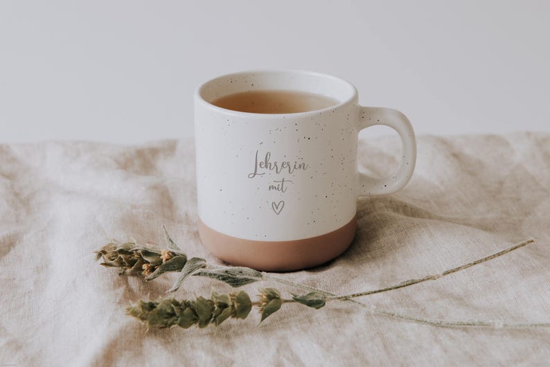 Handgefertigte Keramiktasse mit der Aufschrift "Lehrerin mit Herz" auf einem beigen Leinentuch, umgeben von zarten, getrockneten Blumen. Diese einzigartige Tasse ist das perfekte Geschenk für Lehrerinnen, die mit Liebe und Hingabe unterrichten. Ideal für eine entspannende Teepause und ein schönes Zeichen der Wertschätzung.