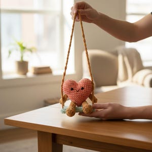 以下が含まれることがあります： 小さなブランコに乗った、笑顔の桃色のハート型の編み人形。人形は茶色の足と腕をしています。ブランコは茶色のロープで吊り下げられており、水色の座席があります。