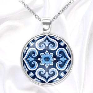May include: A silver-toned pendant necklace featuring a round, blue and white patterned design. The pendant showcases a symmetrical floral motif with swirling details. The necklace hangs on a delicate silver chain, set against a white background.