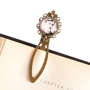 May include: A bronze bookmark with a round glass cameo featuring a black and white photograph of a man. The bookmark has a decorative floral design around the cameo.