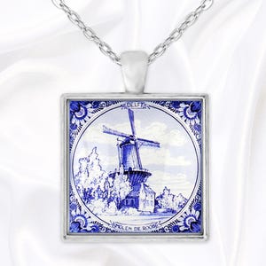 Può includere: Collana con pendente argentato con un ciondolo quadrato. Il ciondolo mostra un mulino a vento di Delft blu e bianco, incorniciato da un bordo floreale. Le parole "DELFT" e "MOLEN DE ROOB" sono visibili.