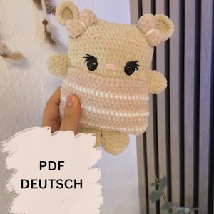 Puede incluir: Un oso de ganchillo beige hecho a mano con una cubierta facial a rayas rosas y blancas. El oso tiene ojos negros y detalles en rosa. La imagen incluye el texto "PDF DEUTSCH" en un cuadro blanco.