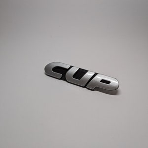 Peut inclure: Un emblème de voiture argenté et noir avec le mot "CUP" en relief.