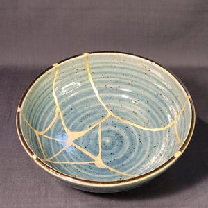 Op de afbeelding: Een ronde, lichtblauwe keramische kom met een donkerbruine rand. De kom heeft een kintsugi-ontwerp, met goudkleurige lijnen die de gerepareerde scheuren benadrukken. De binnenkant heeft een textuur met concentrische cirkels.