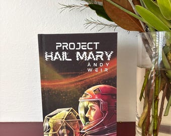 May include: A hardcover book titled Project Hail Mary by Andy Weir featuring a black cover with a space-themed illustration. The artwork depicts a person in a red space helmet looking toward a geometric, golden, crystalline structure in space.
