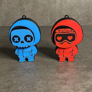 Pode incluir: Duas pequenas figuras estilizadas. Uma é azul com um rosto de caveira e olhos de cruz negra. A outra é vermelha com olhos em forma de coração. Ambas têm capuzes e pequenas argolas no topo para pendurar.