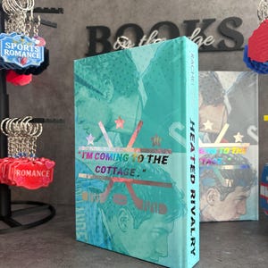 May include: A teal book titled "Heated Rivalry" with a graphic of two men and hockey sticks. The cover features the quote "I'M COMING TO THE COTTAGE." and the author's name, Rachel Reid. Sports romance keychains are in the background.