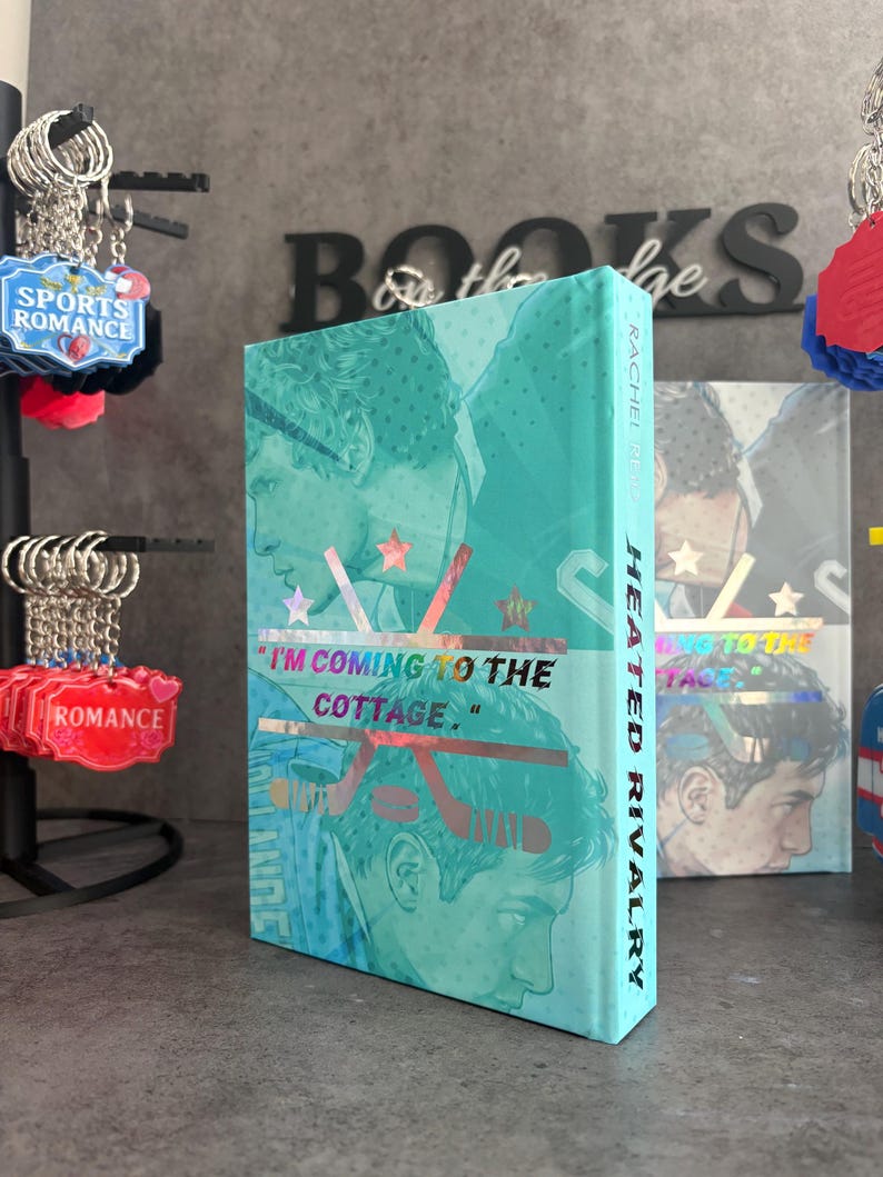 May include: A teal book titled "Heated Rivalry" with a graphic of two men and hockey sticks. The cover features the quote "I'M COMING TO THE COTTAGE." and the author's name, Rachel Reid. Sports romance keychains are in the background.