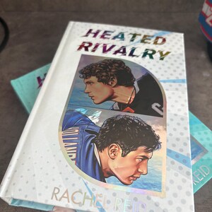 May include: A hardcover book titled "Heated Rivalry" by Rachel Reid. The cover features two men in profile, with a blue and white background. The title is in a rainbow holographic font.