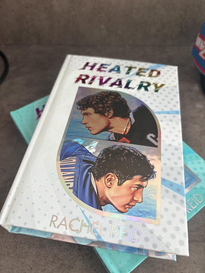 May include: A hardcover book titled "Heated Rivalry" by Rachel Reid. The cover features two men in profile, with a blue and white background. The title is in a rainbow holographic font.