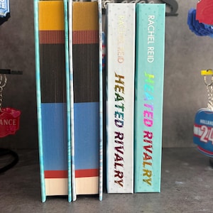 May include: A stack of three books with colorful spines. The books are titled "Heated Rivalry" by Rachel Reid. The spines are white, teal, and blue with gold and pink lettering. The edges of the books are painted in various colors.