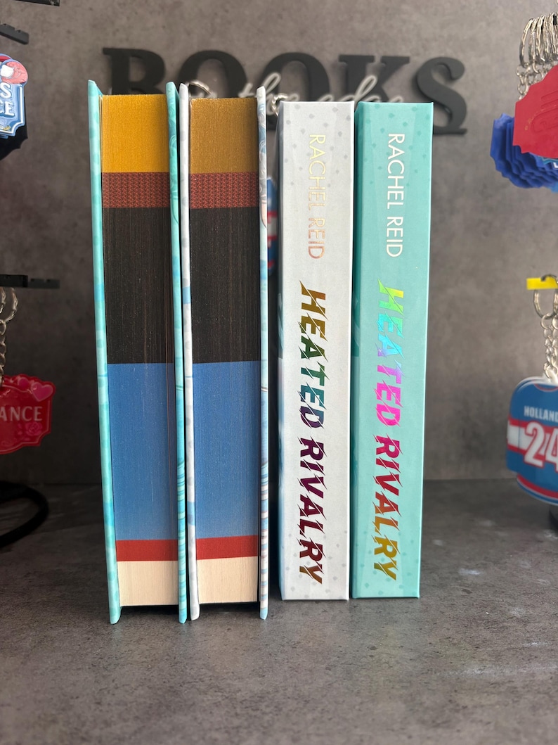 May include: A stack of three books with colorful spines. The books are titled "Heated Rivalry" by Rachel Reid. The spines are white, teal, and blue with gold and pink lettering. The edges of the books are painted in various colors.