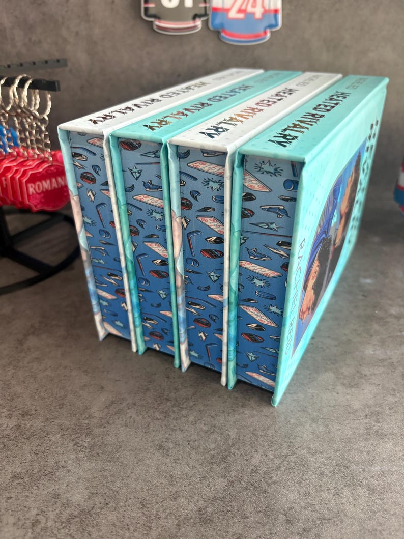 May include: Four books titled "Heated Rivalry" are stacked together. The book spines are white with teal text. The book covers are teal with a pattern of sports equipment. The book's edges are teal.