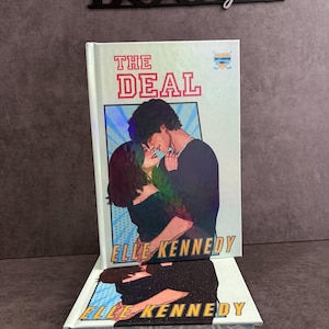 Op de afbeelding: Een hardcover boek getiteld The Deal van Elle Kennedy met een holografische coverillustratie van een omhelzend stel. Het ontwerp bevat een achtergrond in stripstijl, dikke rode tekst en een klein hockey-embleem. De onderkant toont muzikale noten.